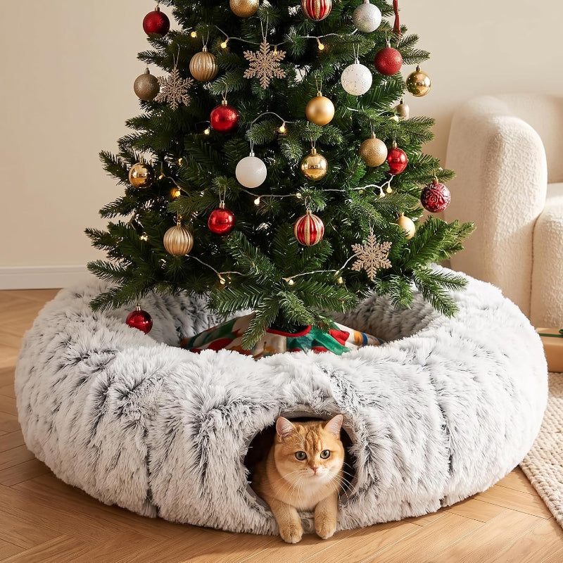 A fluffy round pet bed in light gray faux fur sits under a decorated Christmas tree on a wooden floor, with an orange cat peeking out from the front opening.