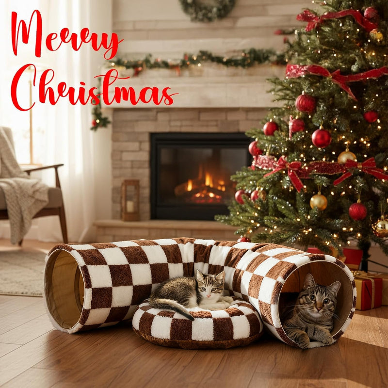 Two cats in a checkered pet bed with a Christmas tree and fireplace in the background.