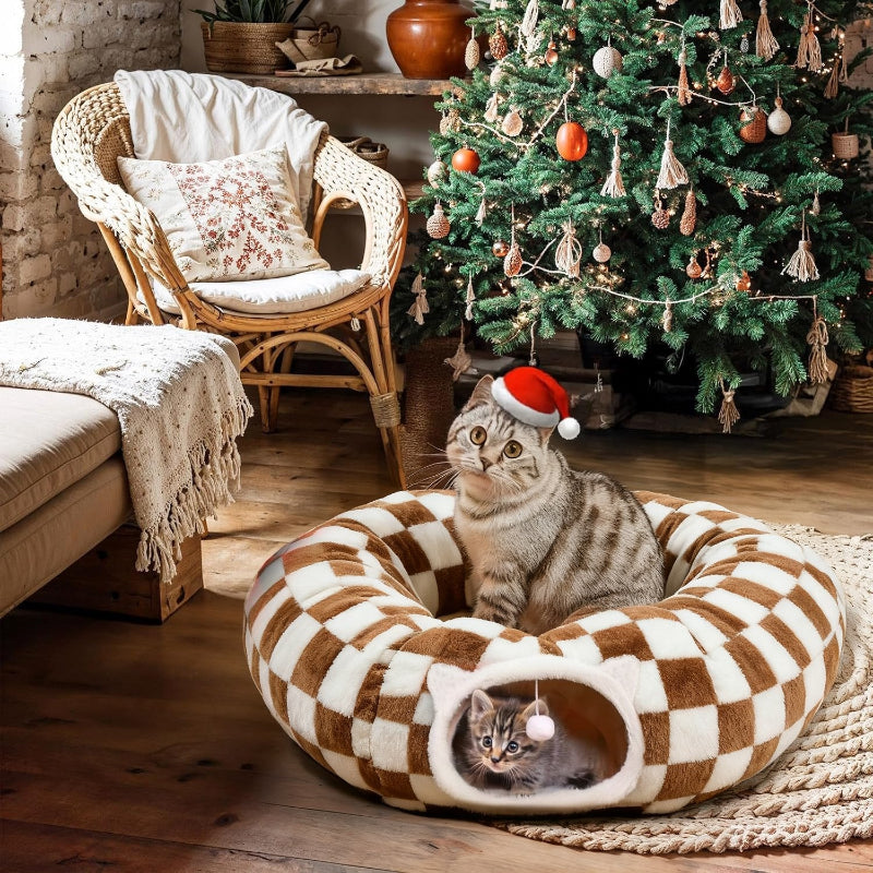 Cat wearing a Santa hat sitting on a checkered pet bed in a cozy room with a Christmas tree.