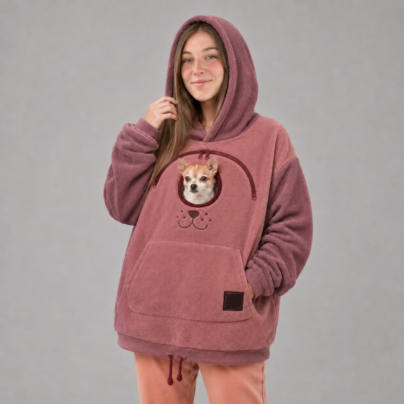 A woman wearing a dusty rose fleece hoodie with a built-in dog pocket featuring a Chihuahua graphic, standing against a solid gray studio background.