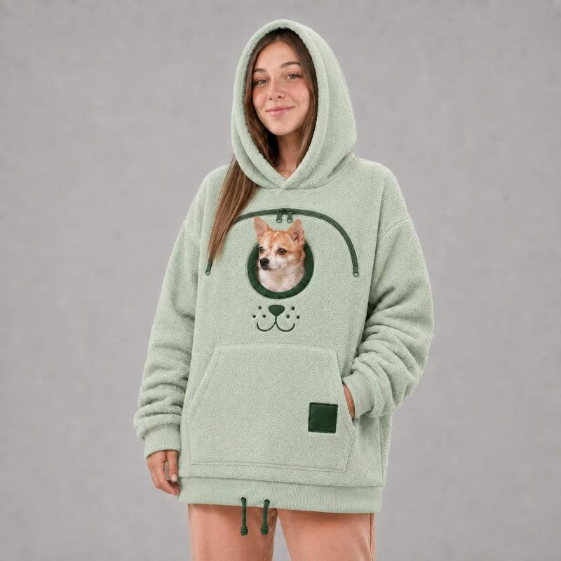A woman wearing a light sage green fleece hoodie with a graphic of a Shiba Inu dog in a circular space helmet on the front, paired with peach pants, against a neutral gray background.