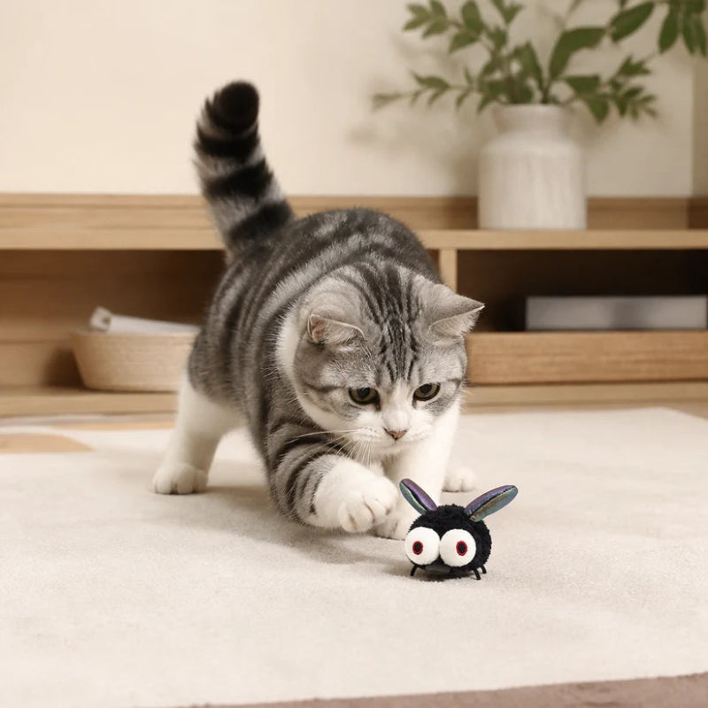 A gray and white tabby cat stretches to investigate a small black bunny toy with large white eyes on a light beige rug in a cozy indoor setting.
